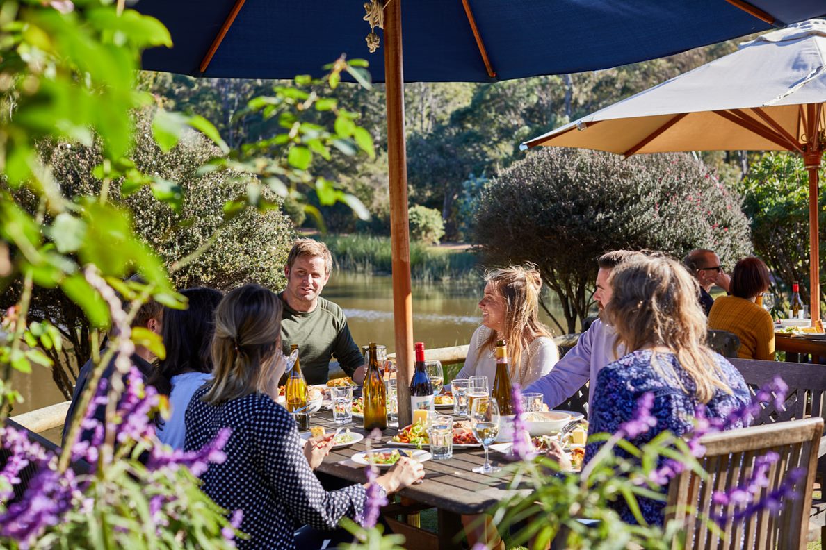 A group sharing a meal and wine at a sunny outdoor table overlooking a pond