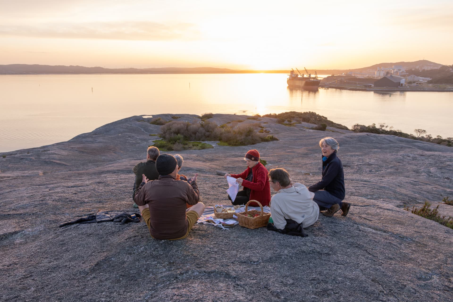 A small group gathered at a bush picnic table beneath trees at sunset, with a guide and warm evening light