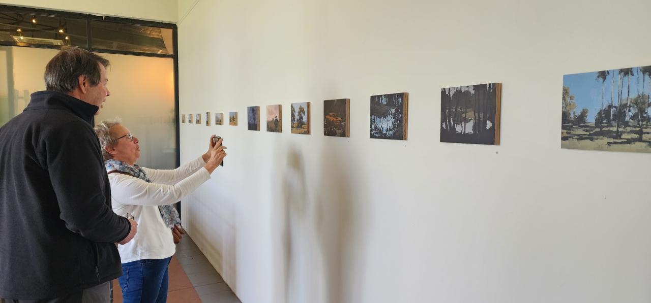 A man and a woman viewing small landscape paintings on the white wall of a gallery
